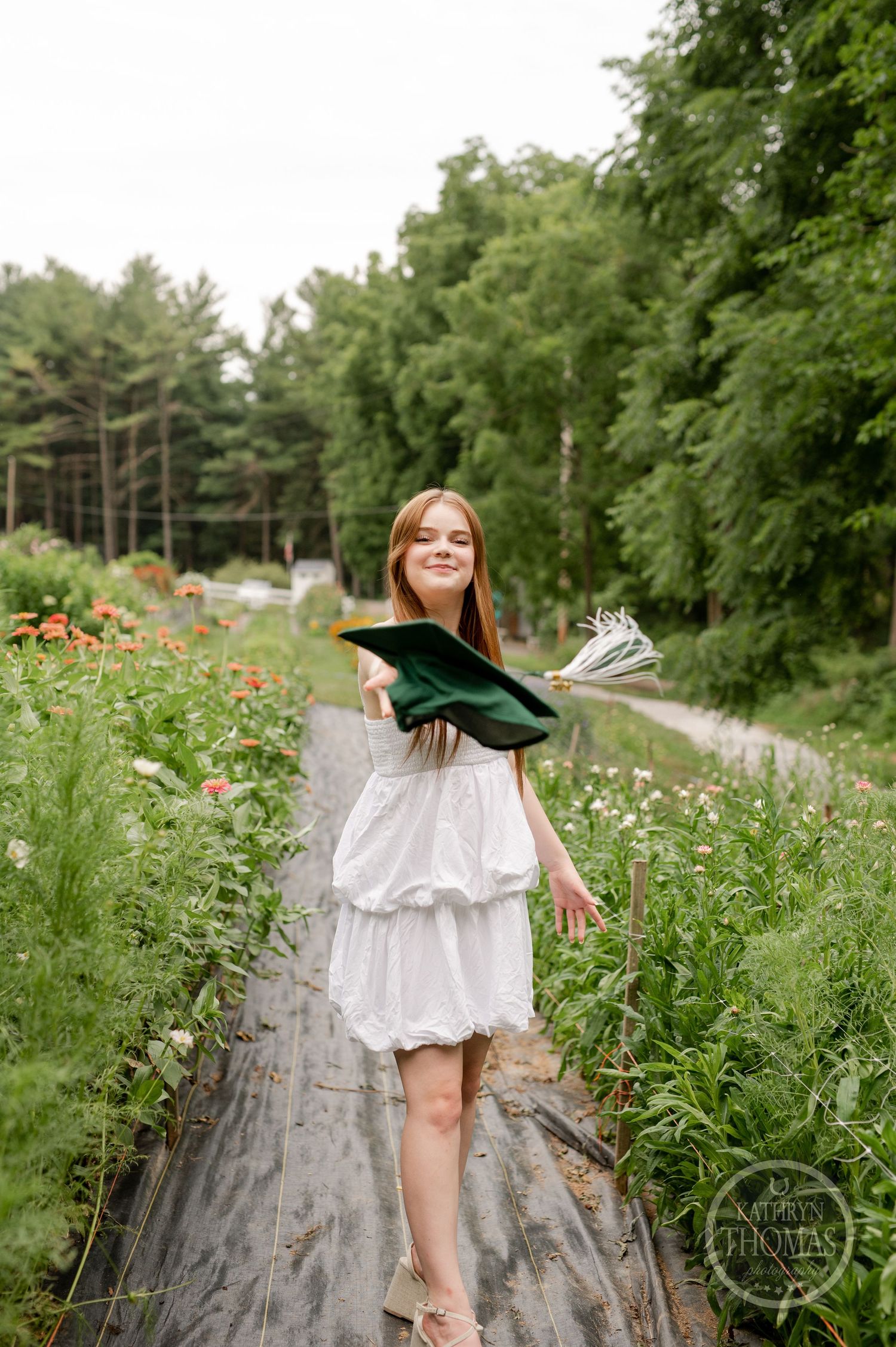 Graduate in white dress poses with diploma on wooden boardwalk surrounded by wildflowers.