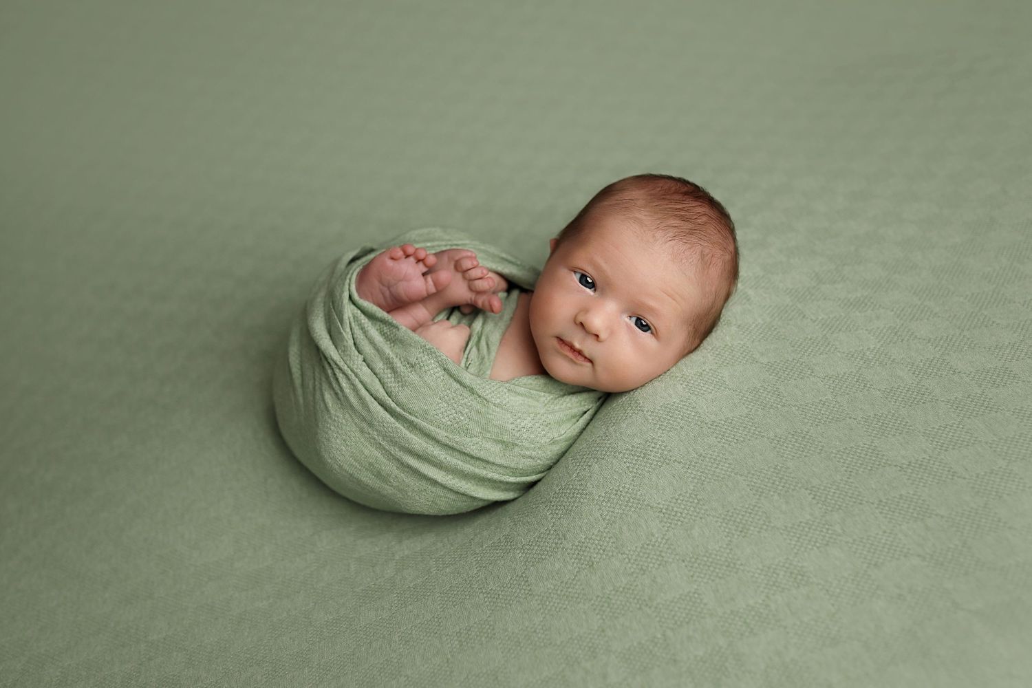 Newborn swaddled in soft mint green fabric lying on matching background during professional photo session.