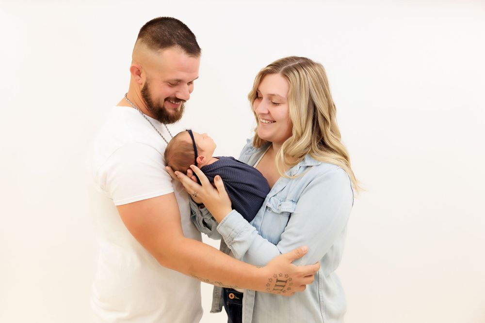 A couple gazes lovingly at their newborn baby wrapped in dark clothing against a white background.