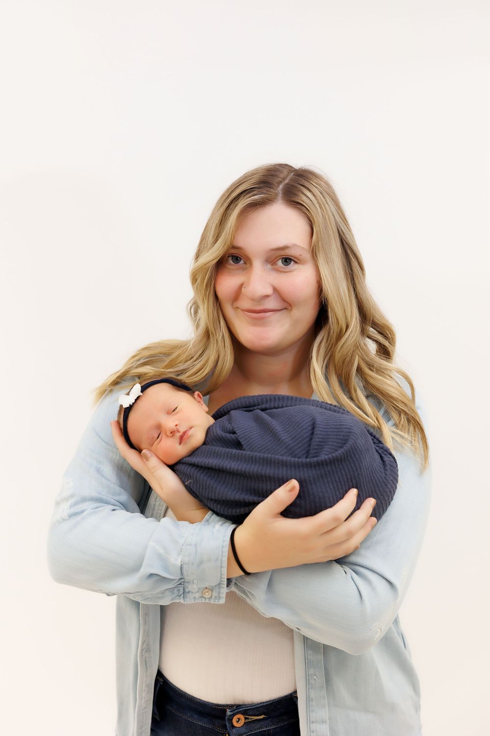 Parent in light denim jacket holds sleeping newborn while smiling against white background.
