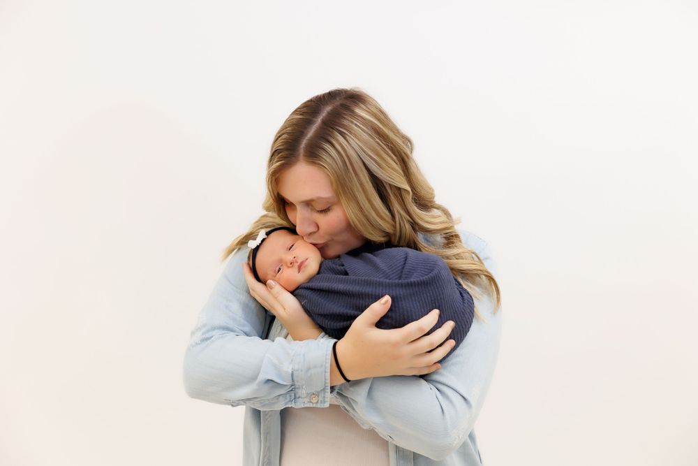 Parent tenderly embraces sleeping newborn wrapped in navy swaddle against white backdrop.
