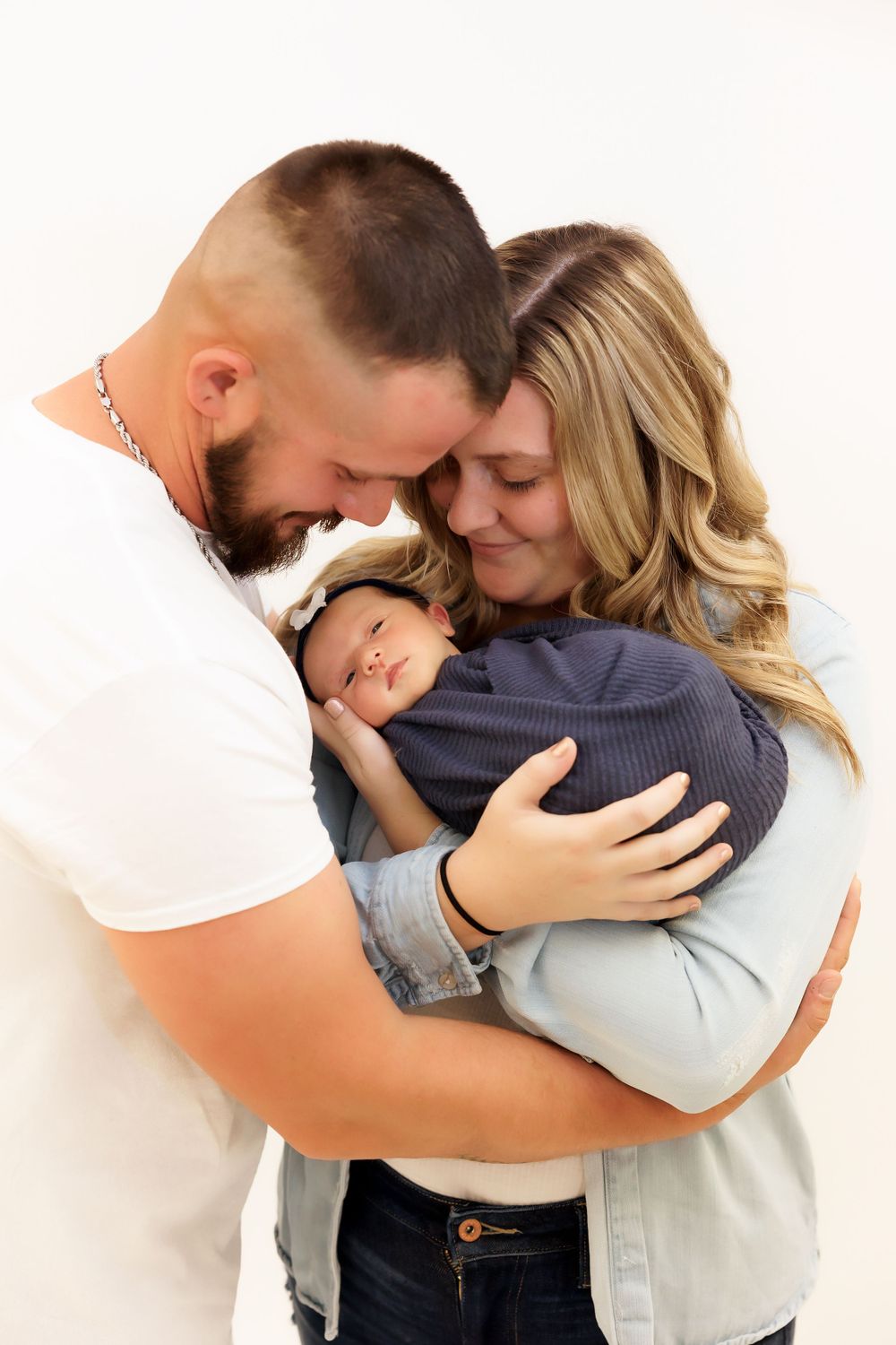 An intimate family portrait showing parents snuggling their newborn baby in a navy wrap.