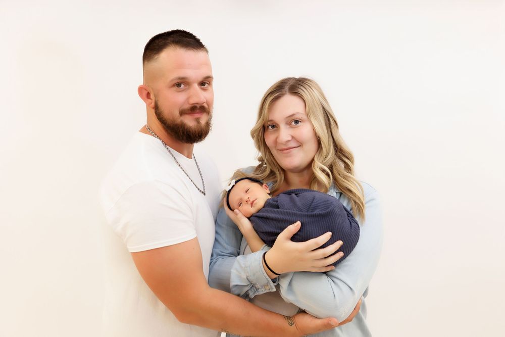 A couple in white and gray casual clothing holds a sleeping newborn wearing navy blue against a light background.