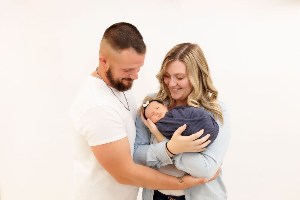 Couple in white and casual attire tenderly embracing their newborn baby against a light background.