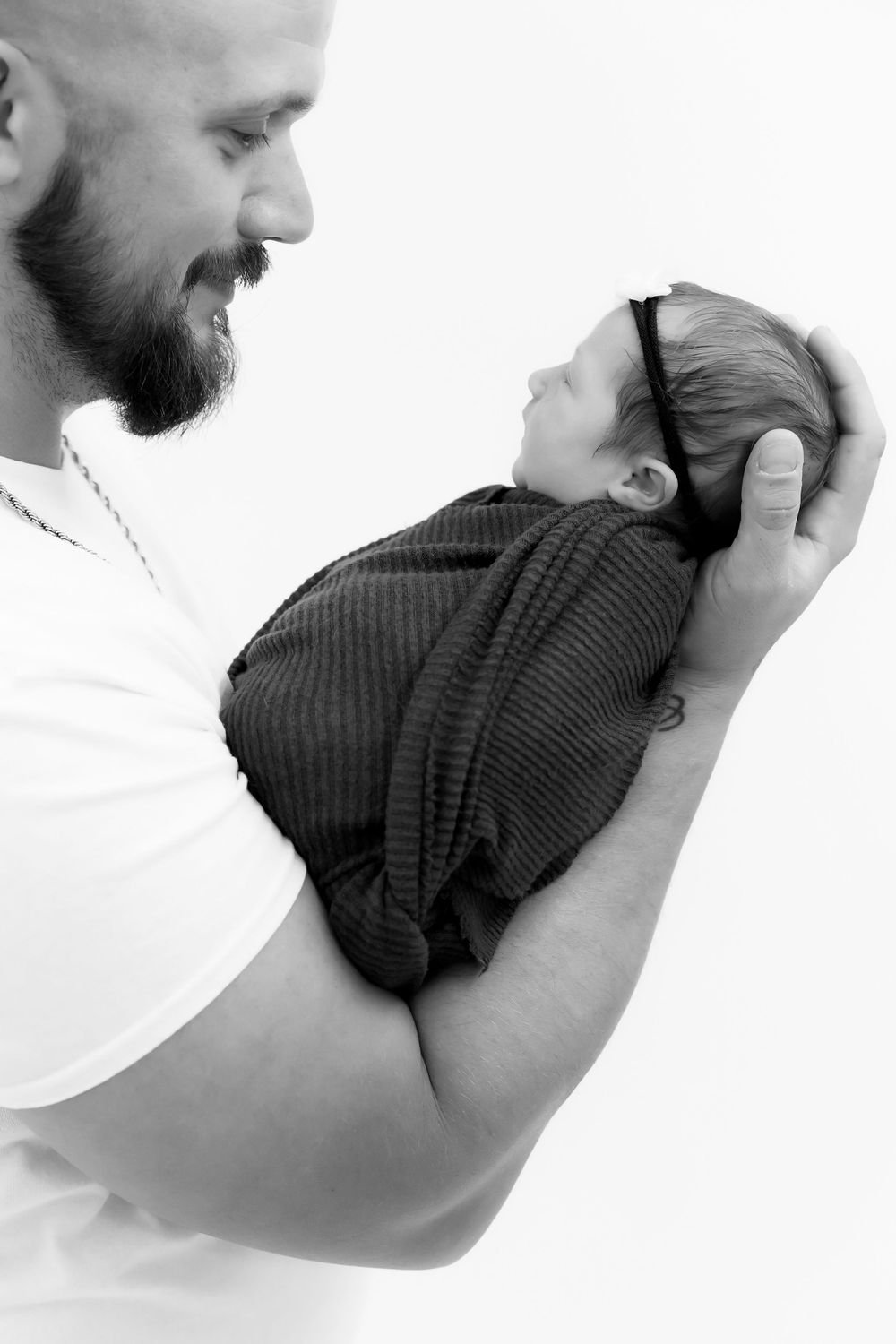 Black and white portrait of a bearded parent lovingly holding a swaddled newborn baby against white background.