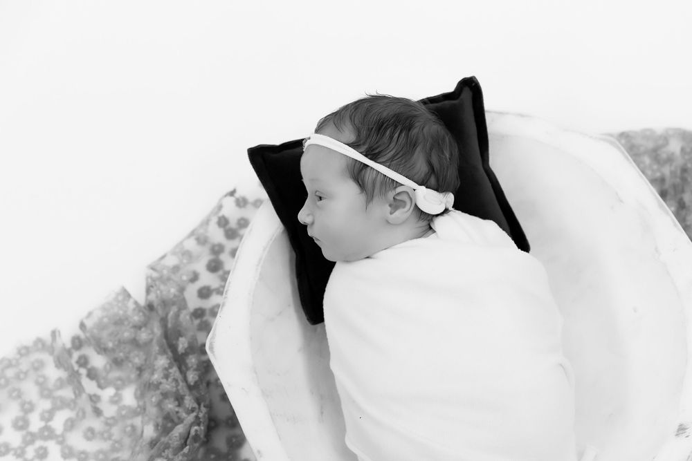 A sleeping newborn wearing a bow lies on soft white bedding in black and white.