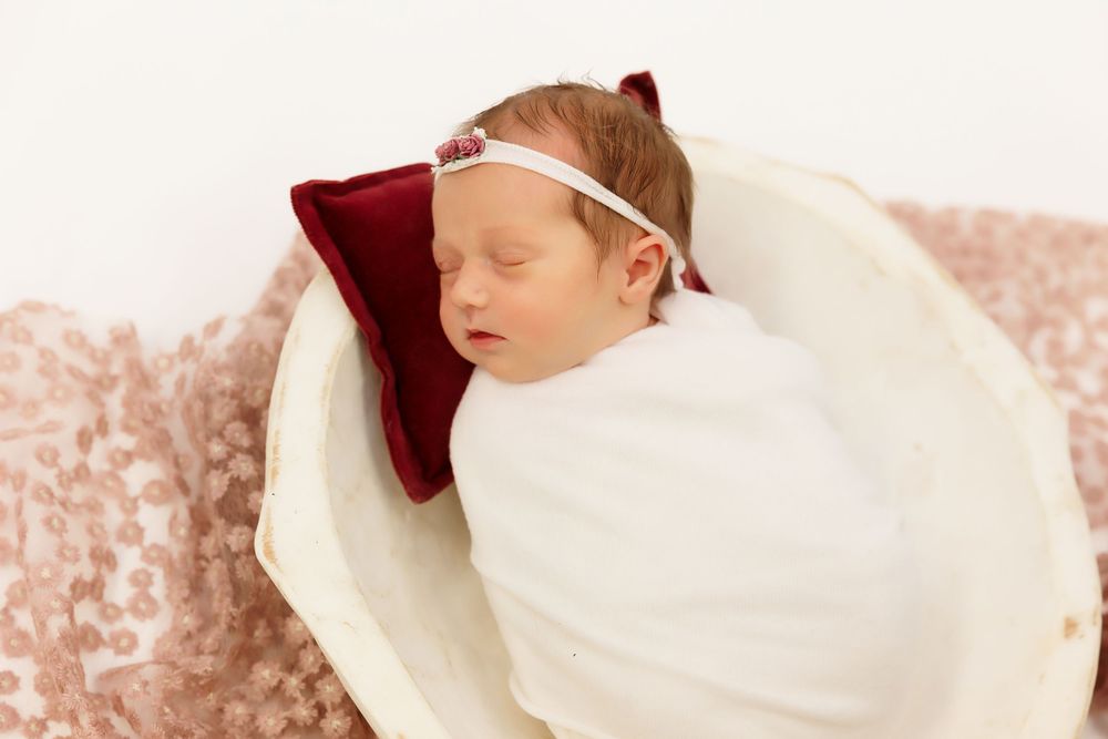 Sleeping newborn wrapped in white blanket with red bow headband in cream basket.