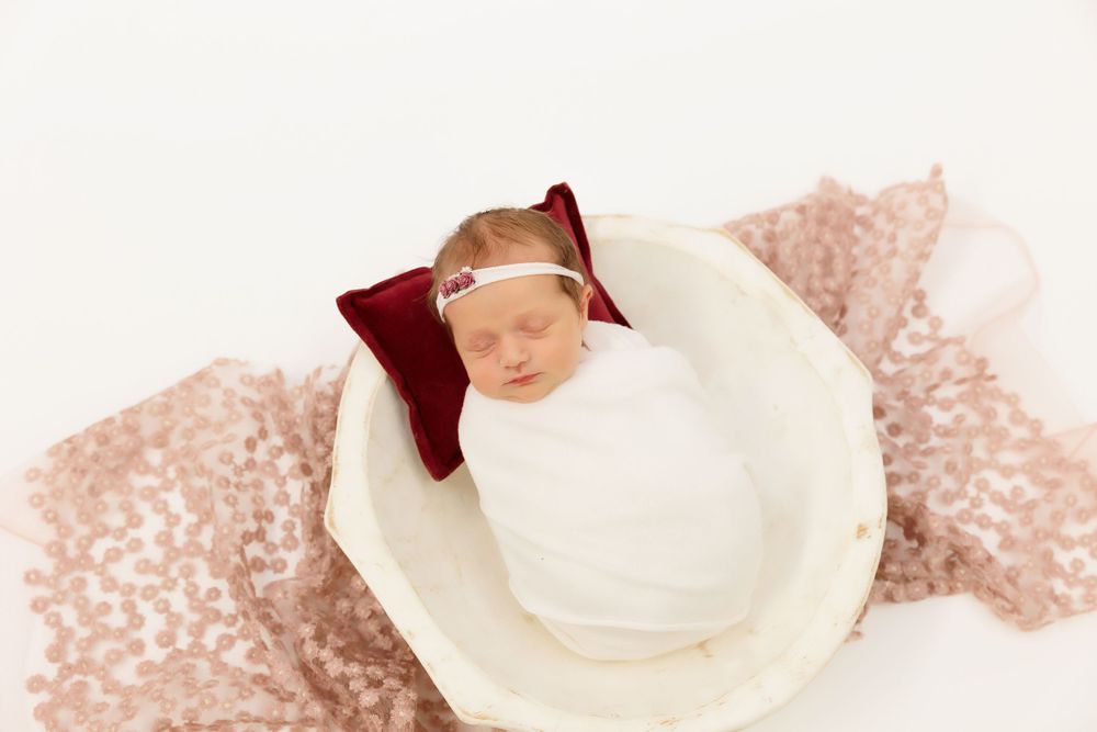 Newborn sleeps peacefully in white bowl with burgundy bow on sparkly rose gold backdrop.