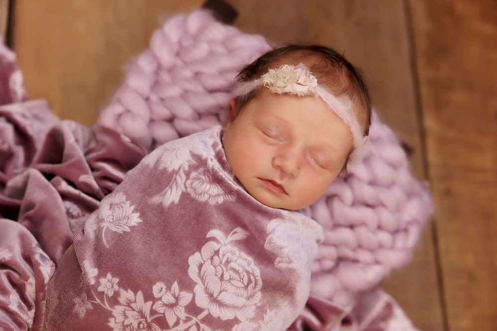 Sleeping newborn wearing lace headband on pink textured blanket with floral print wrap.