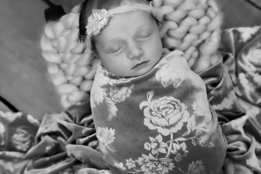 Black and white portrait of sleeping newborn with floral wrap and lace headband on knit blanket.