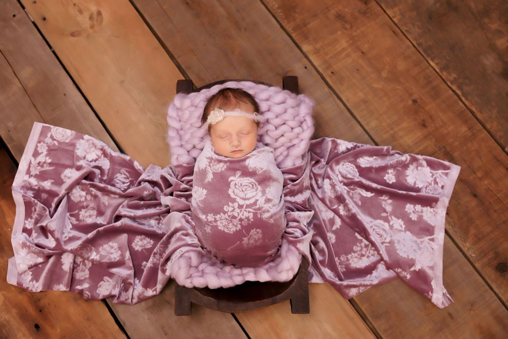 Newborn baby resting on wooden floor surrounded by pink floral fabric and plush texture.