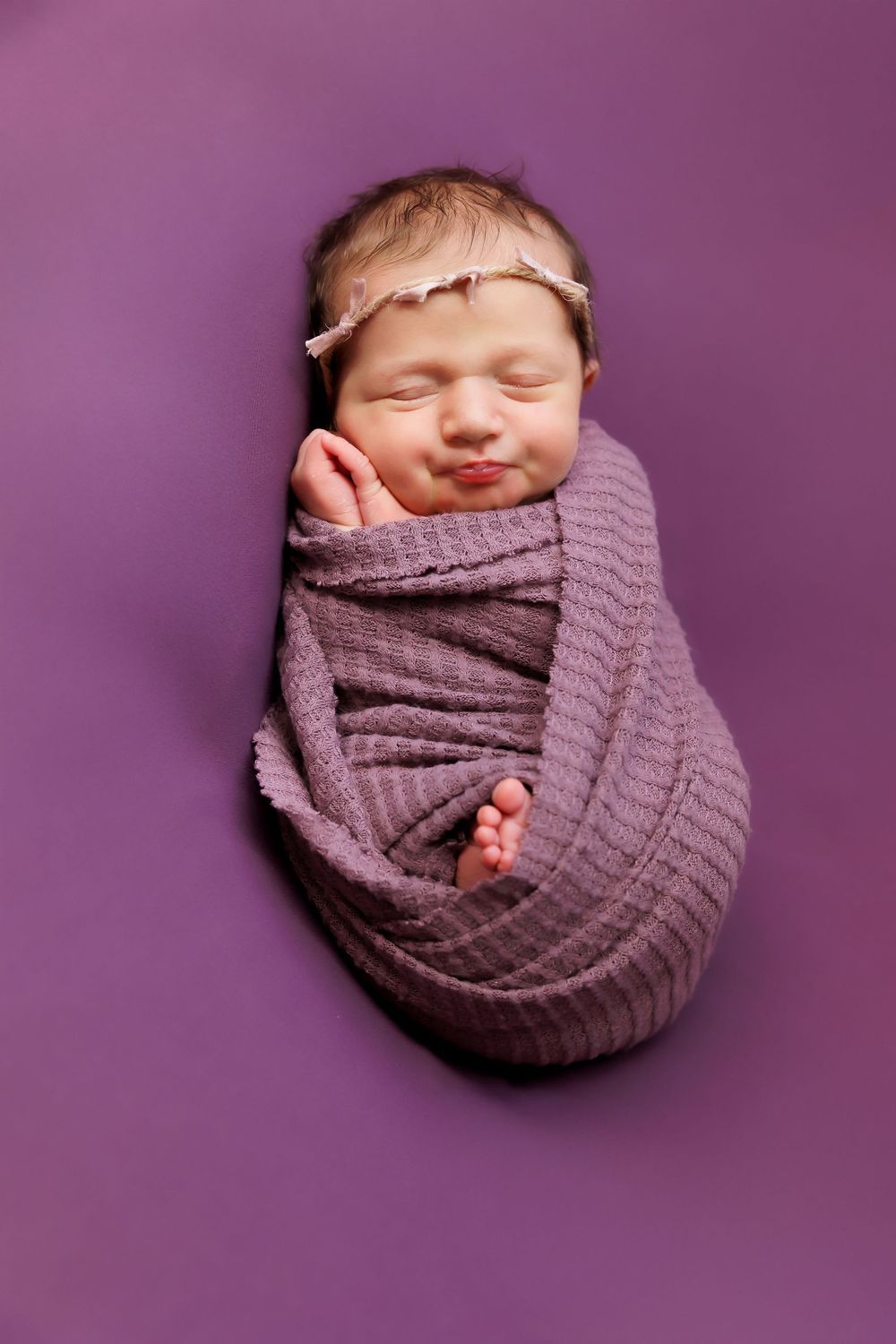 Newborn baby sleeping peacefully wrapped in purple knit blanket on purple background.