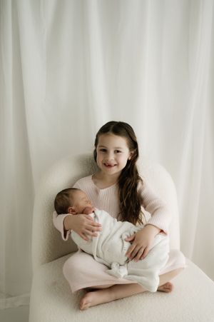 Two children sitting together in white chair against light backdrop in minimalist portrait setting.