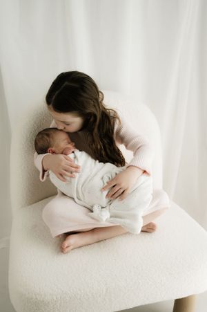 Intimate portrait of tender moment with baby in white outfit sitting on cozy white chair against neutral background.
