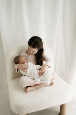 Gentle motherhood moment captured in soft white tones as woman holds sleeping baby in cozy armchair.
