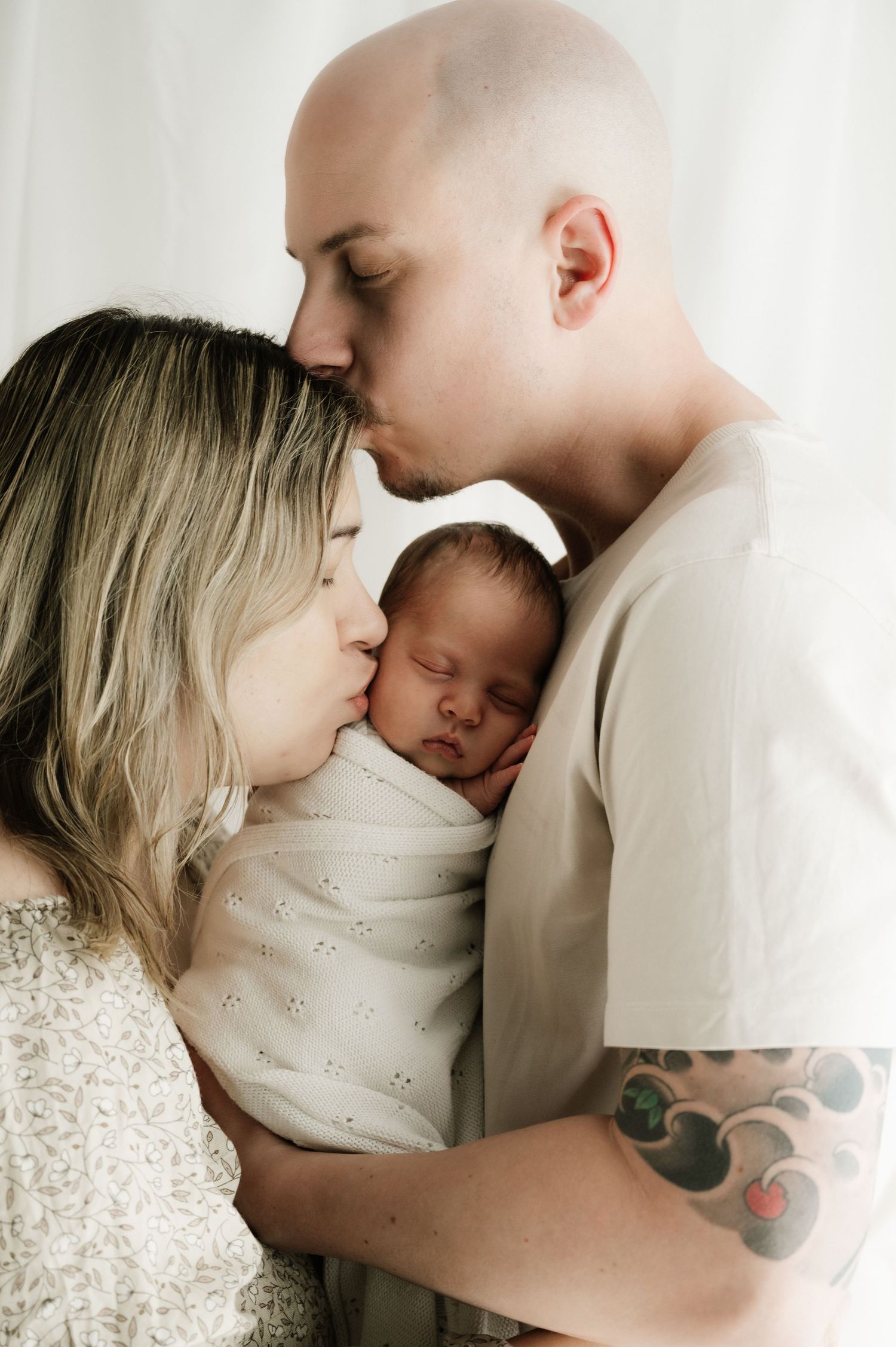 A bald man with tattoos and woman in white embrace their sleeping newborn baby in an intimate portrait.