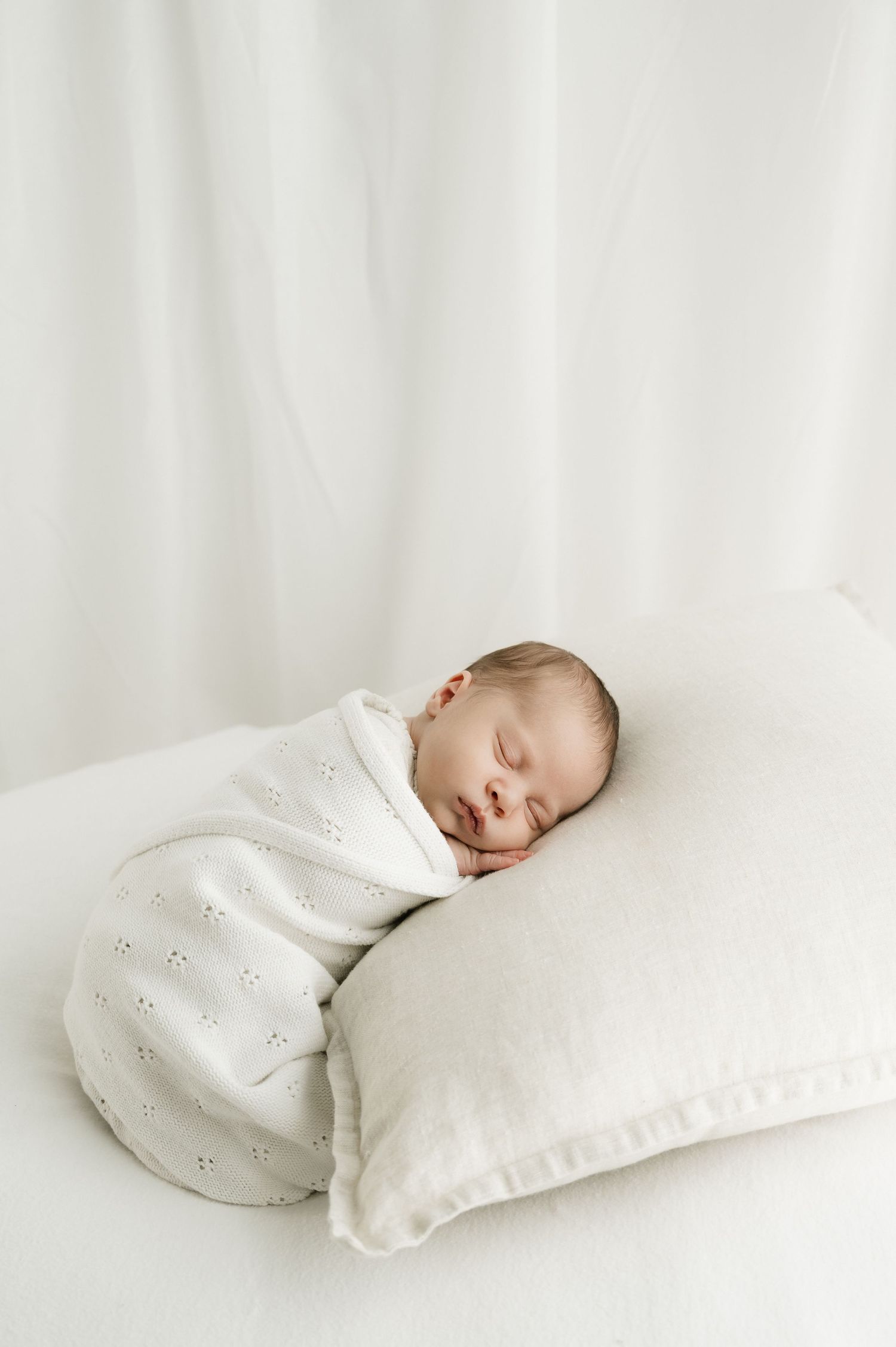 Sleeping newborn baby wrapped in white knit blanket resting on light colored pillow against white background.