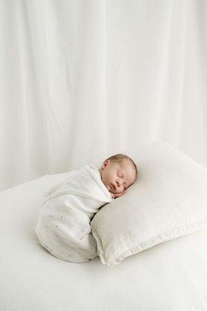 A peaceful sleeping baby wrapped in white swaddle rests on a white pillow against light fabric backdrop.