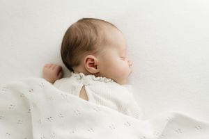 A sleeping baby lies peacefully on white textured bedding in a soft, side profile view.
