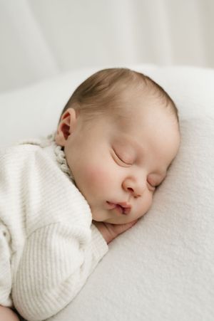A sleeping baby wearing a white sweater rests peacefully on soft white bedding.