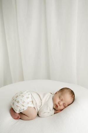 A peaceful sleeping baby wearing white lies curled up on a white backdrop with soft fabric draping behind.