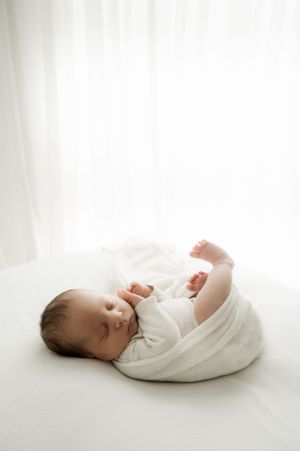 Sleeping newborn baby wrapped in white swaddle blanket lying on light backdrop in peaceful studio setting.
