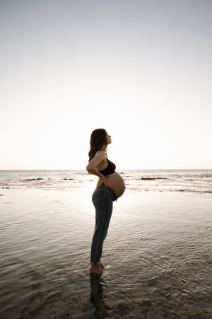 A silhouetted pregnant figure stands at the edge of calm ocean waters during sunset, arching back in a serene pose.
