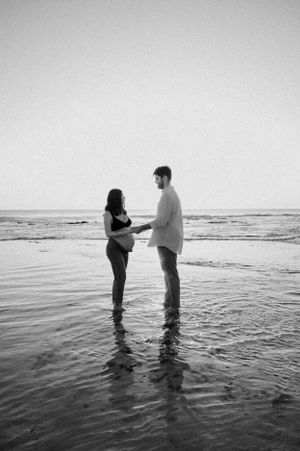 A couple stands together in shallow ocean waves at the beach in a romantic black and white photograph.