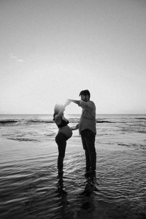 A couple dancing in shallow ocean water at sunset, captured in black and white photography.