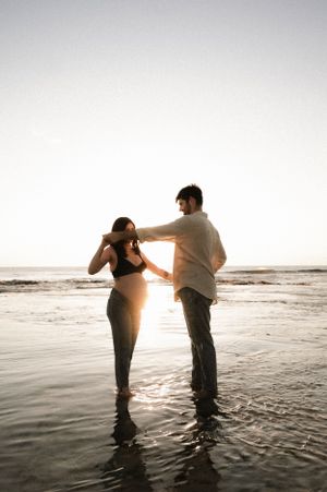 A couple stands together in shallow ocean water at sunset, creating a romantic silhouette against the bright sky.