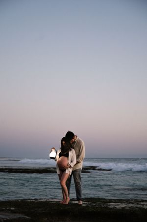 Silhouette of a couple sharing an intimate moment on rocky shore at dusk with ocean waves in the background.