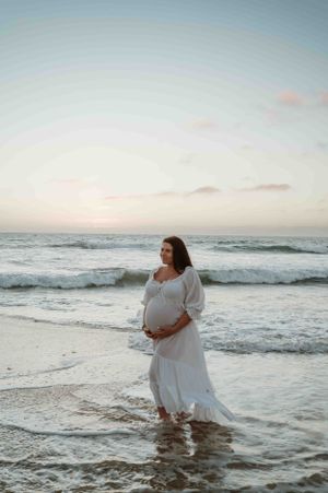 Ethereal maternity photo of a woman in a white dress standing in ocean waves at sunset.