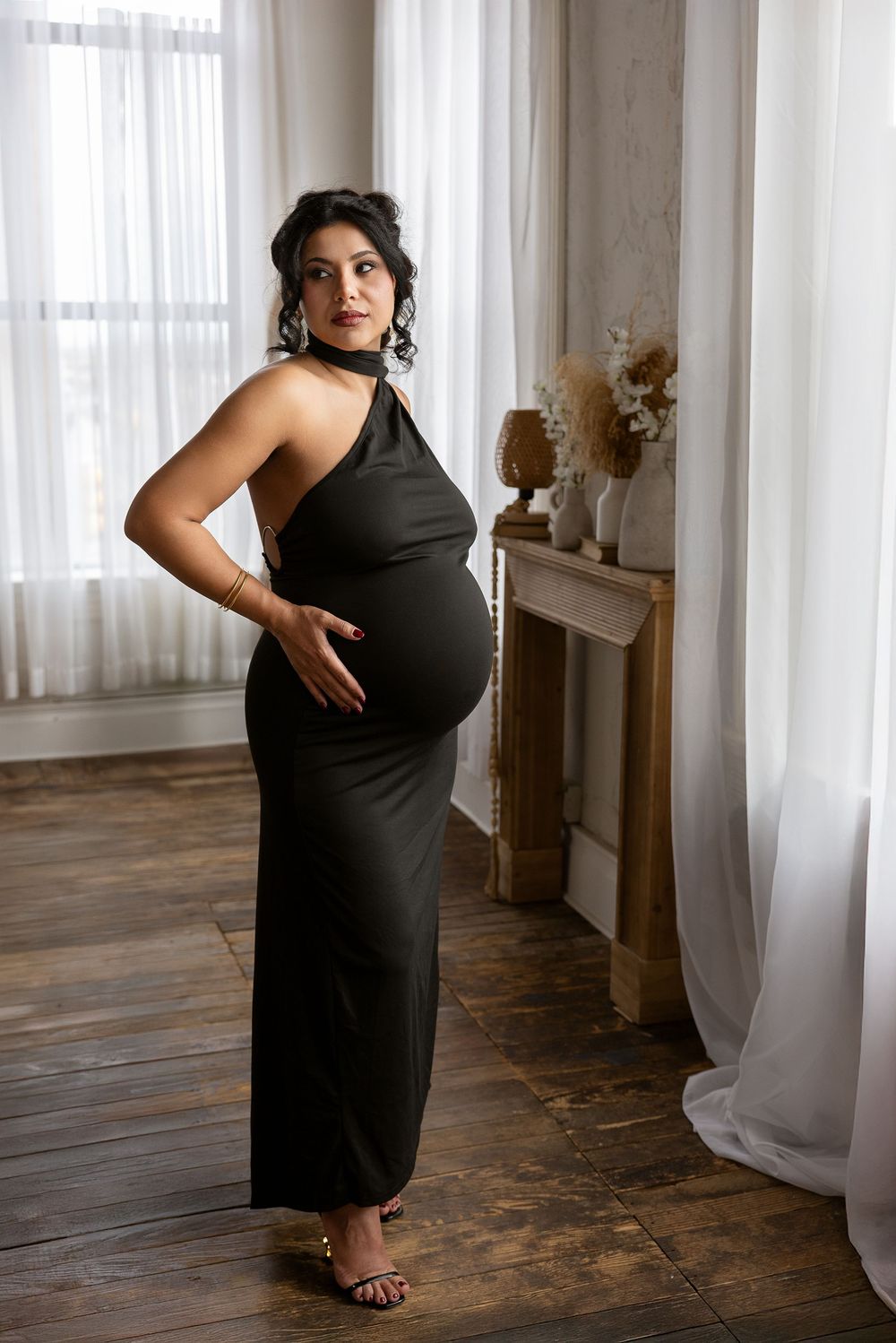 Elegant maternity photoshoot featuring a long black halter dress against white curtains and wooden floors in natural light.