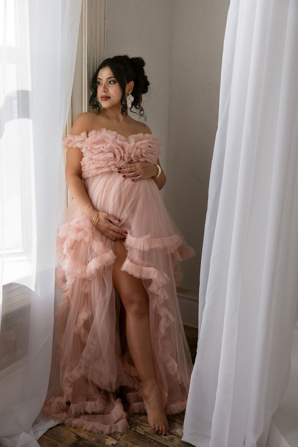 Pregnant woman in flowing blush pink tulle gown poses elegantly by window with sheer white curtains.