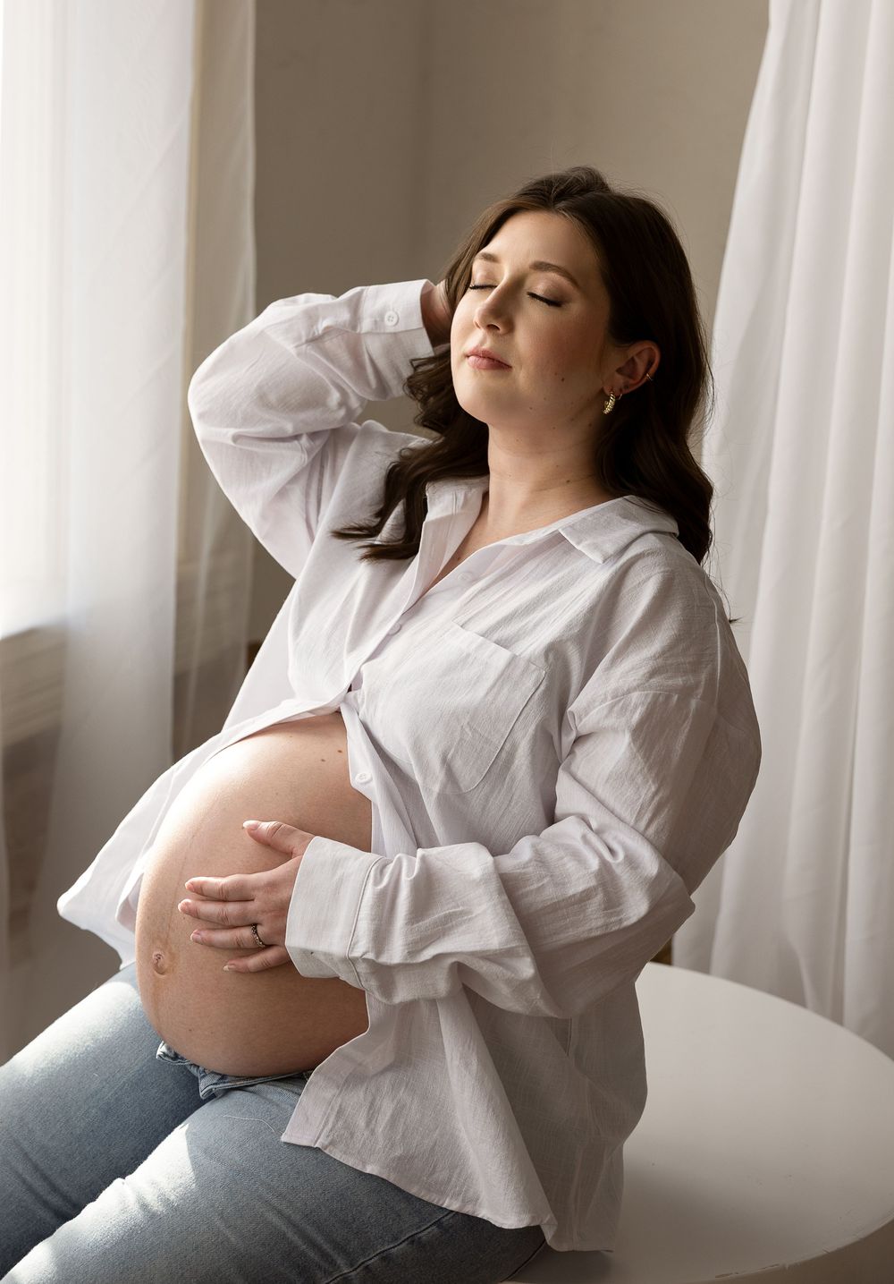 A pregnant woman in a white shirt reclines against a light background in a series of artistic maternity photos.