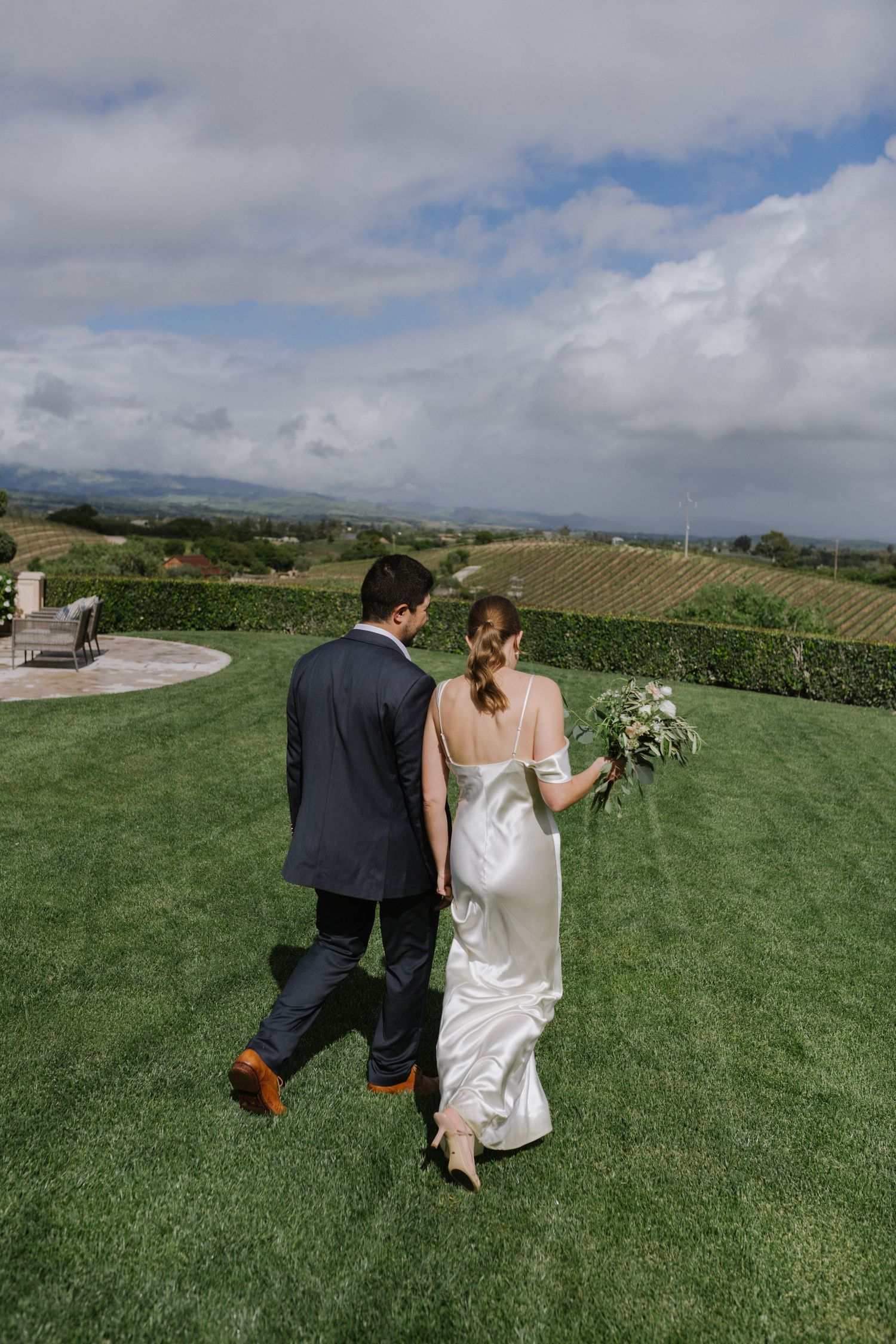 Couple walking on manicured lawn with dramatic cloud-filled sky and rolling hills in background.