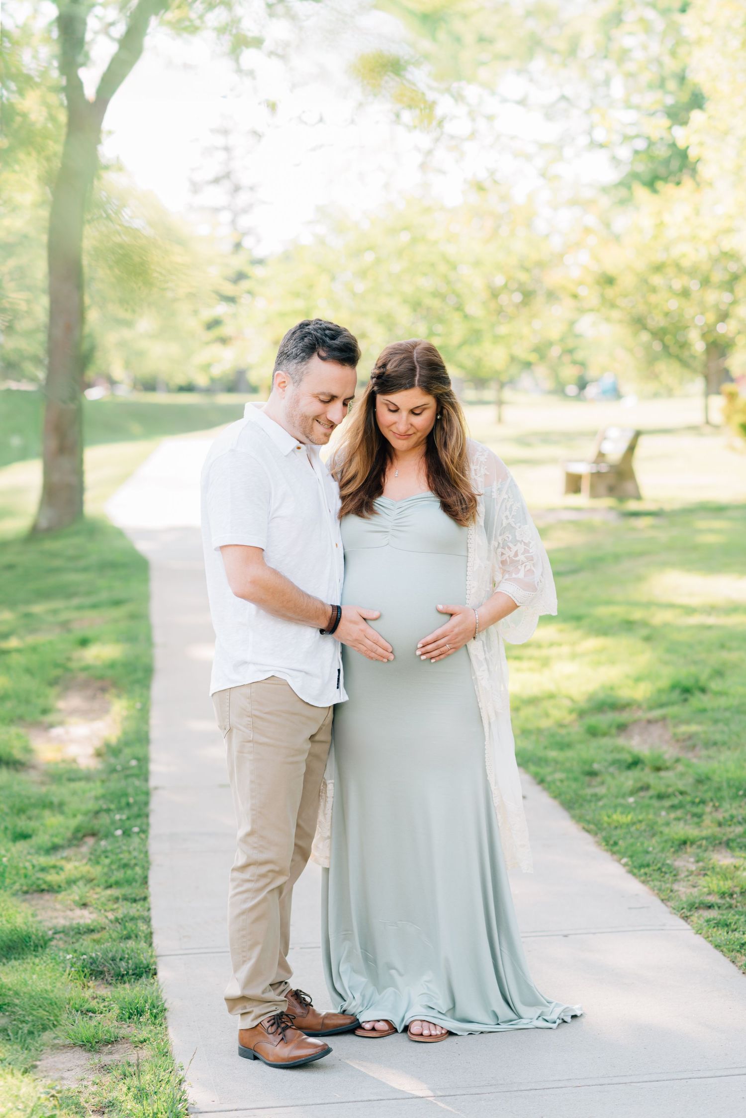 A couple standing on a sidewalk near a lake look down at her pregnant belly.