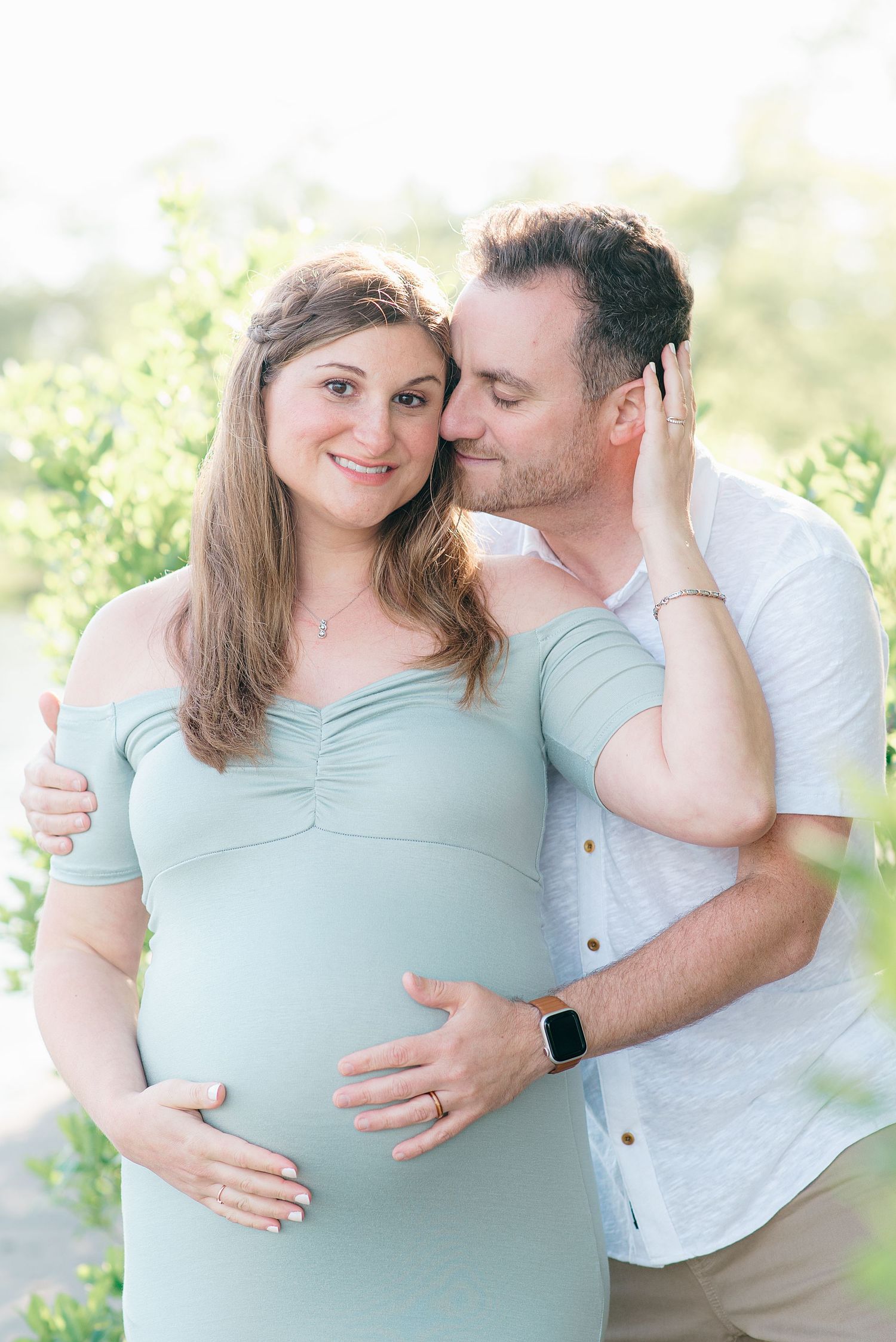 A woman in a light blue dress holds her partner's face and smiles during a maternity session in Monmouth County nj.