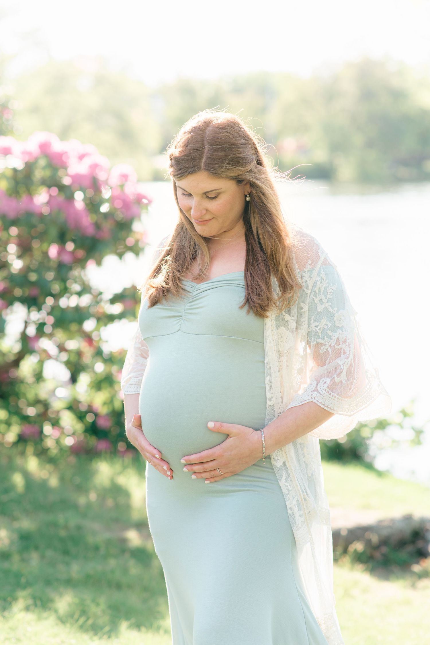 An expecting woman holds her belly and looks at it in front of a lake with flowers behind her.