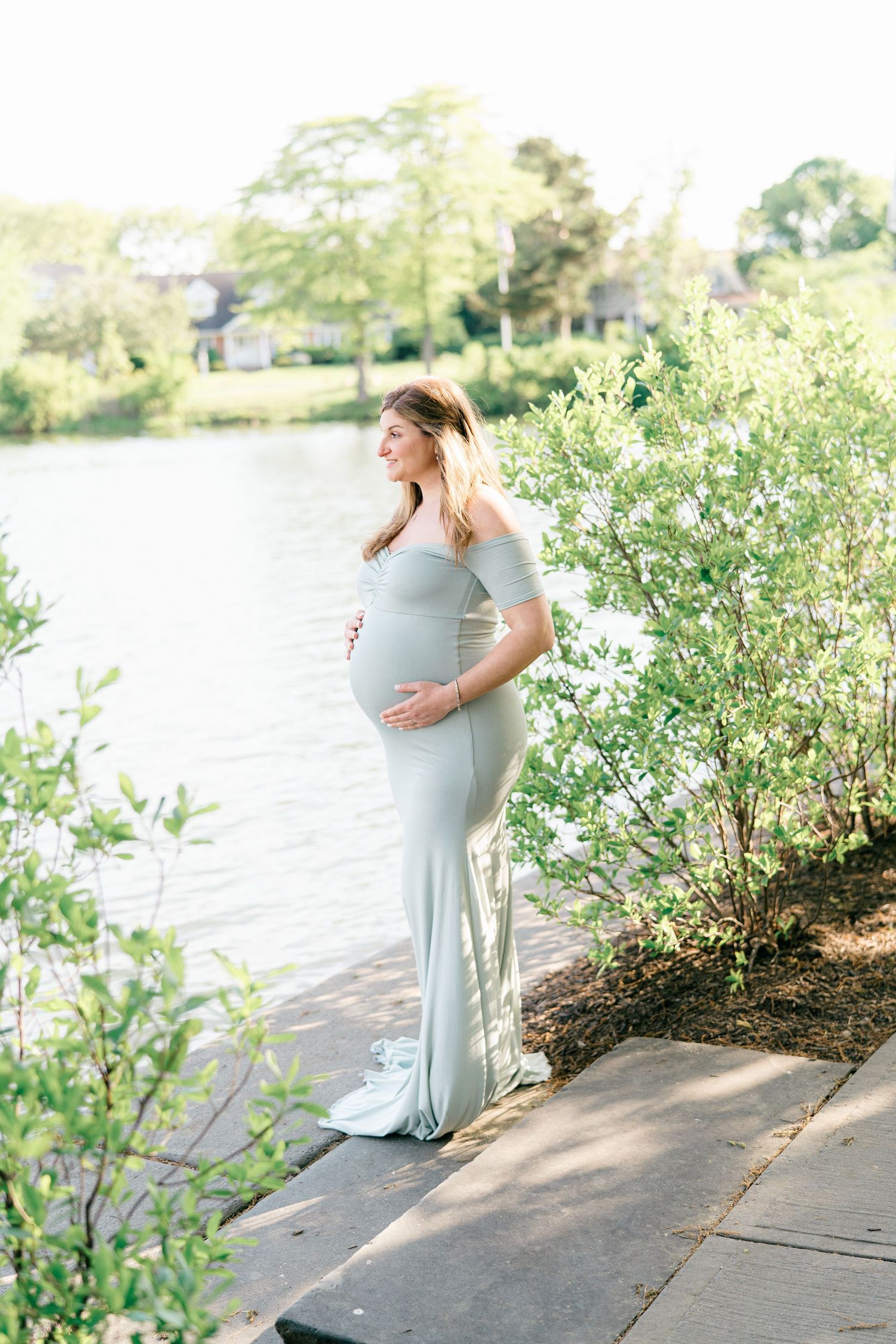 Woman in tight blue maternity dress looks out over a lake in New Jersey.