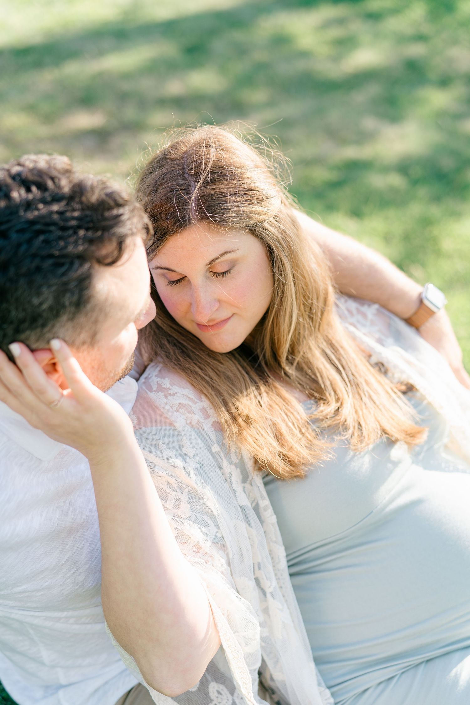 A woman in a blue dress gently touches her husbands face while sitting on the grass.