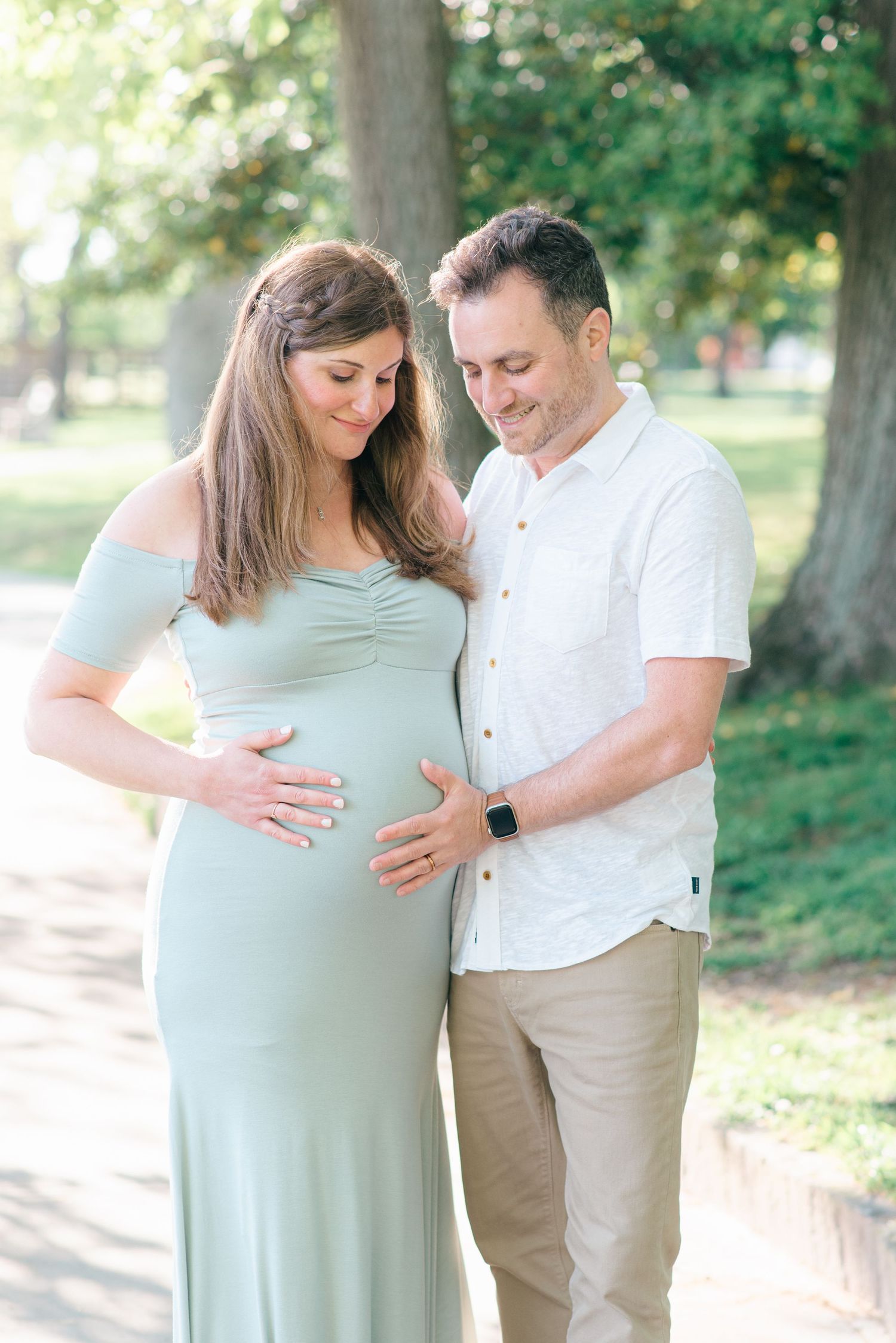 Mom-to-be and man in a white shirt and tan pants standing near a lake with trees and greenery behind them.