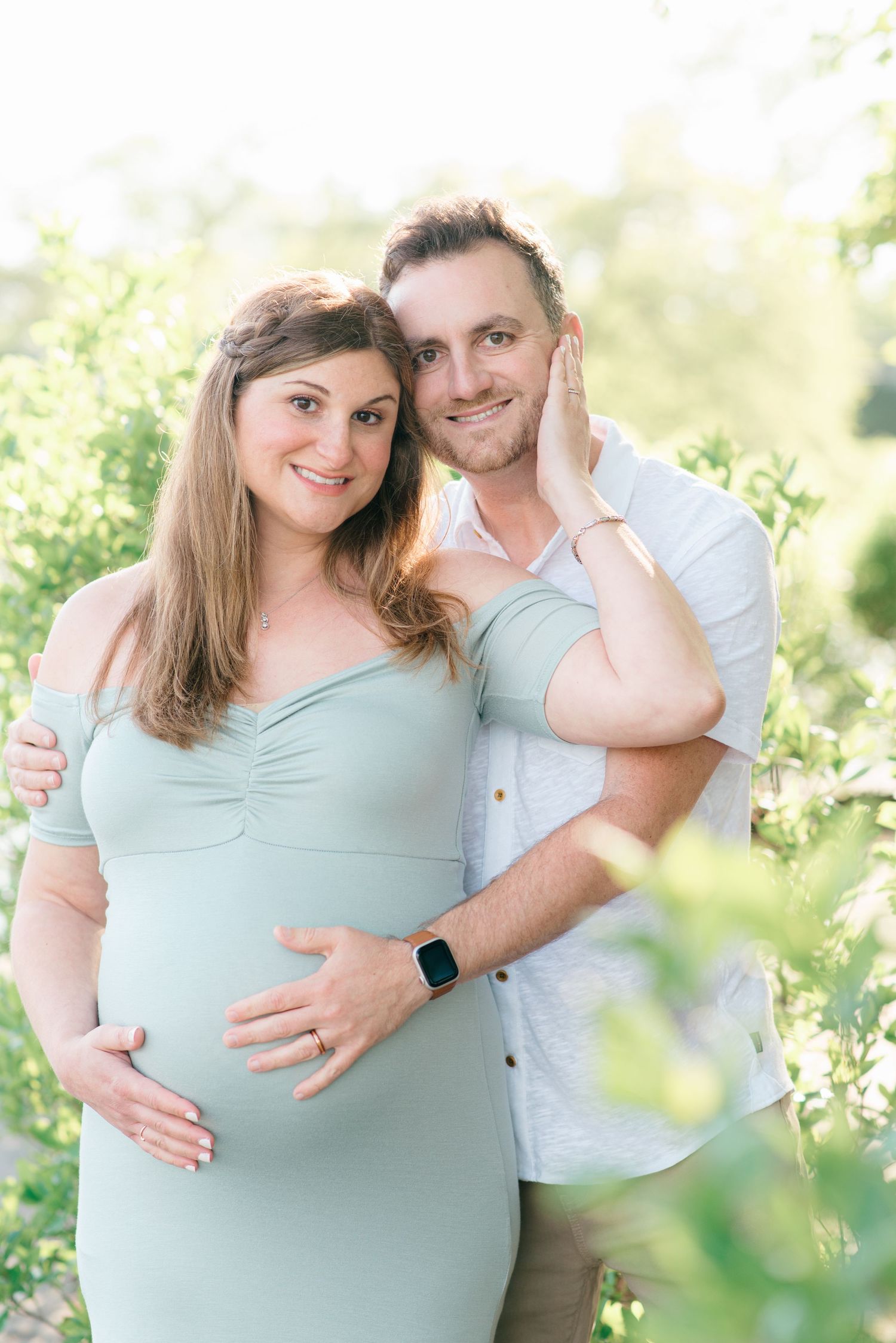 Mother to be holds her husband's face while posing surrounded by greenery.
