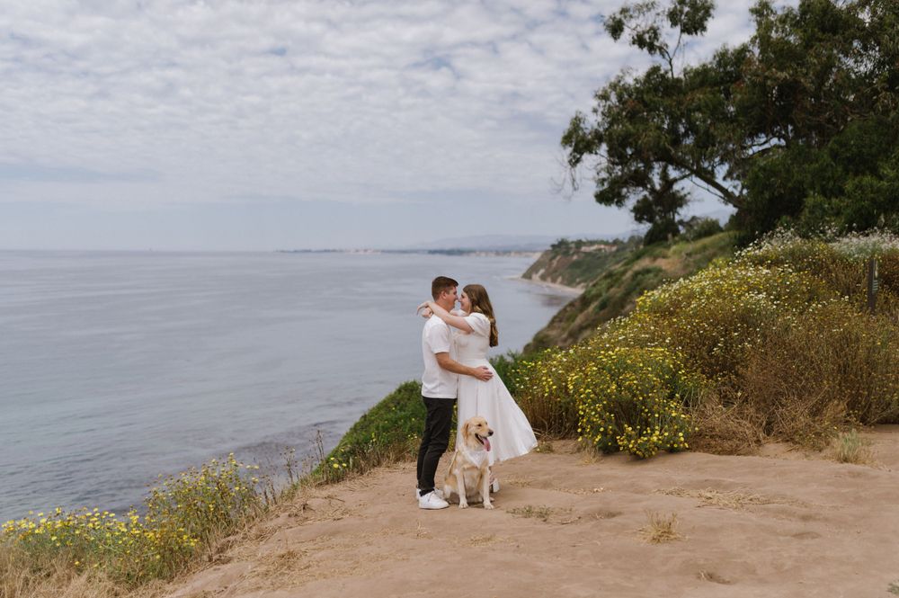 Couple with their dog sharing an embrace on a coastal cliff overlooking the ocean.