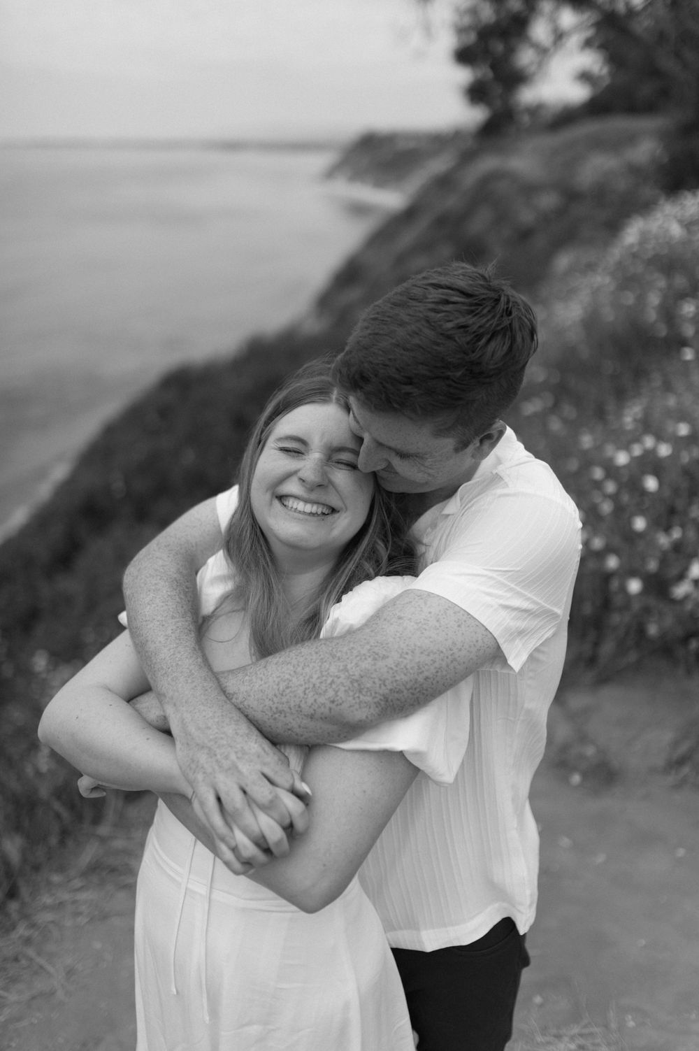 An intimate black and white portrait of a couple embracing on a coastal cliff.