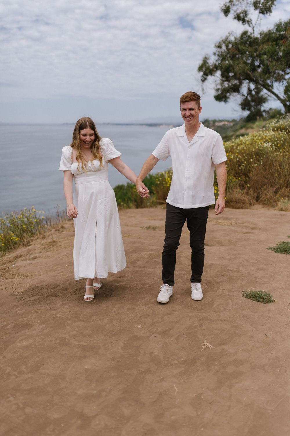 A couple stands hand in hand on a coastal path overlooking the ocean on a sunny day.