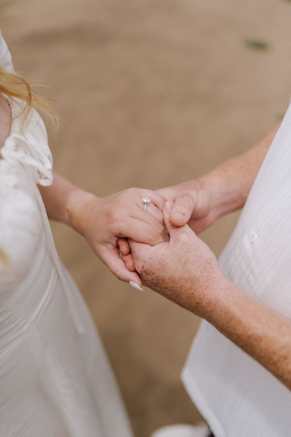 Close-up of hands tenderly clasped together, showcasing engagement rings against white attire.