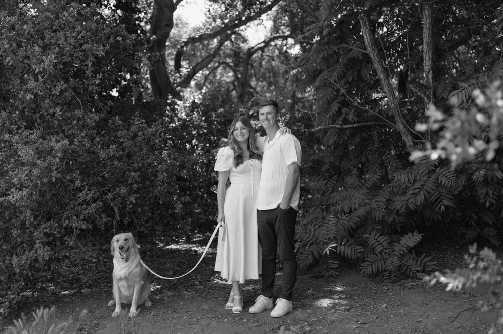 A couple in white outfits stands outdoors with their golden retriever dog in a black and white photograph.
