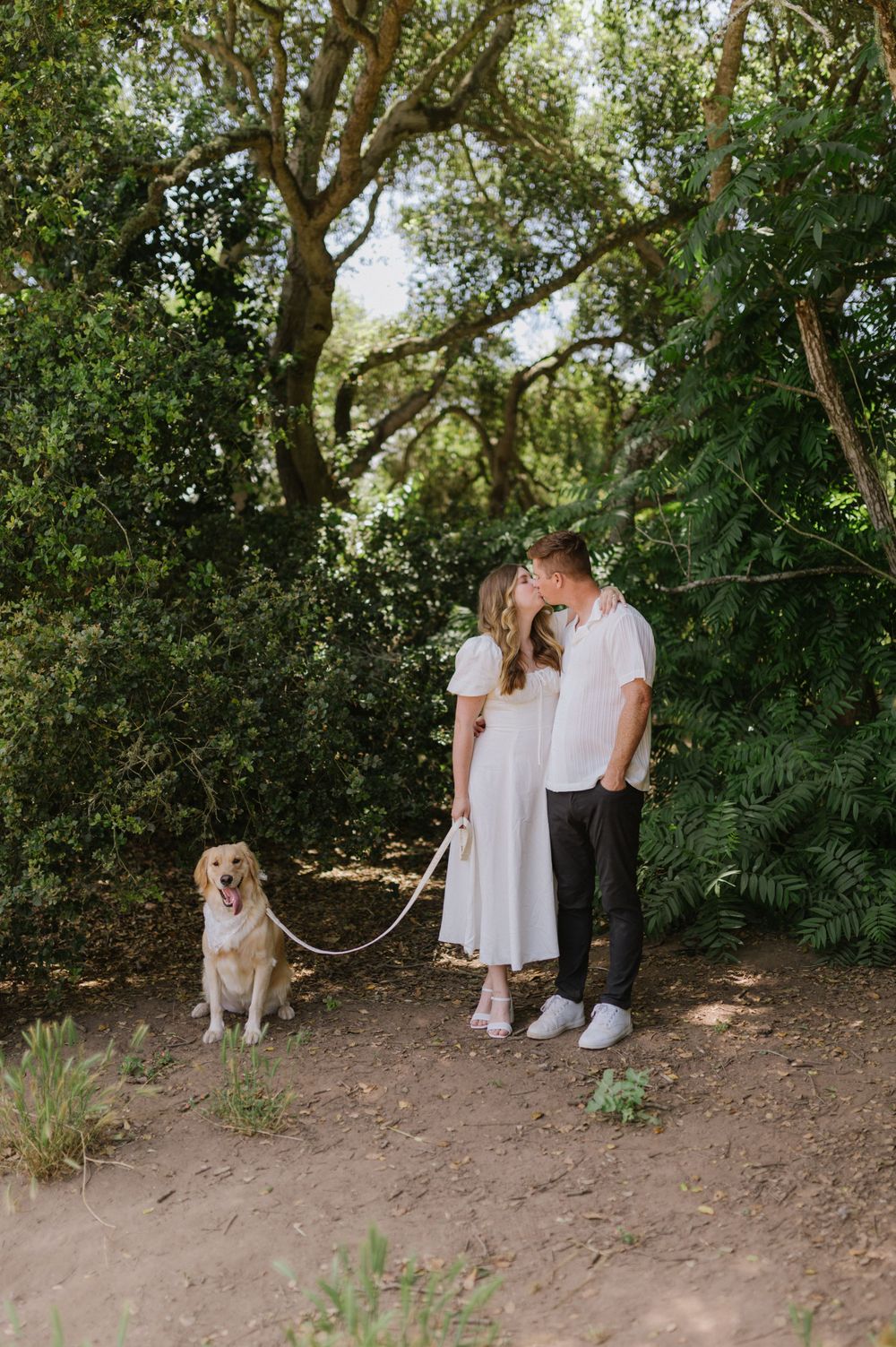 A couple in white clothing stands on a nature trail with their happy golden retriever on a leash.