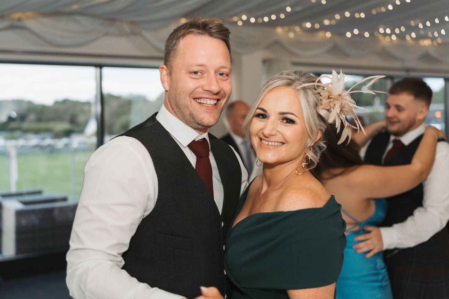 Smiling couple dressed in formal attire dance together under string lights at an evening celebration event.